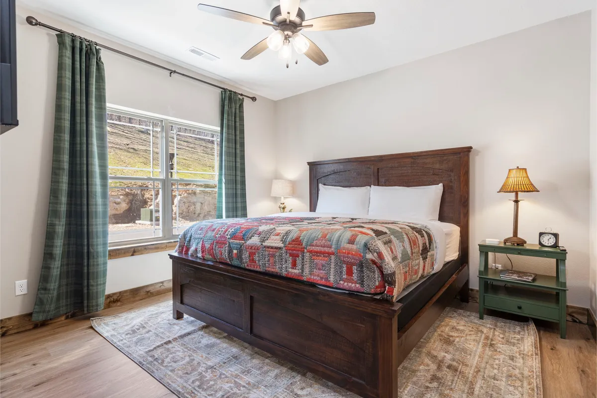 A tranquil bedroom featuring a comfortable bed with a soft coverlet, a large window draped with curtains that allows natural light to stream in, a wall-mounted TV for entertainment, and wooden doors suggesting a private ensuite bathroom nearby. The décor is cozy and peaceful, emphasizing relaxation.