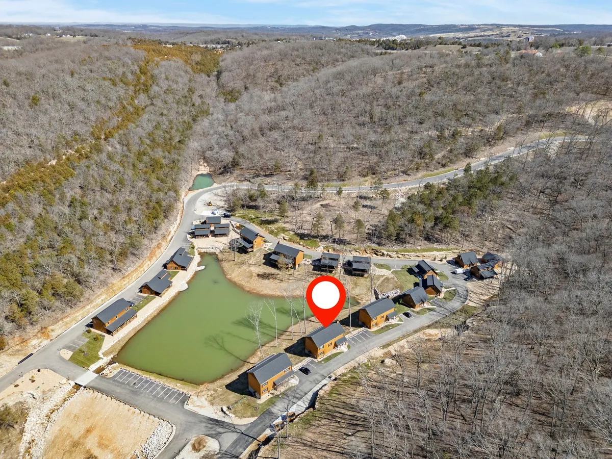 This image depicts a tranquil scene featuring cabins arranged around a serene pond, with gentle hills and natural landscaping in the background. The setting exudes peacefulness and is surrounded by lush greenery, providing a sense of escape into nature.