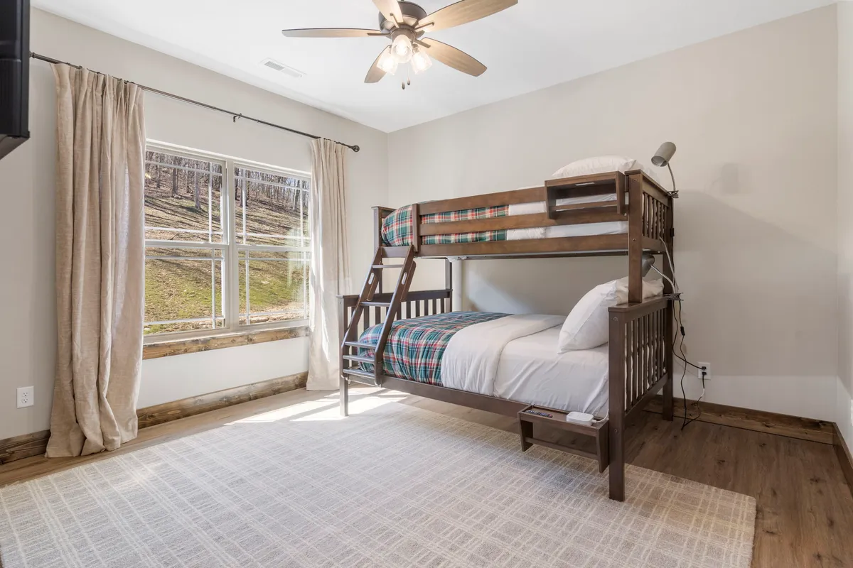 A cheerful bunk room with sturdy wooden bunk beds, a large window illuminating the space, and a textured rug on the floor. The room radiates playfulness and warmth, clearly designed for younger guests to enjoy both restful nights and playful moments.