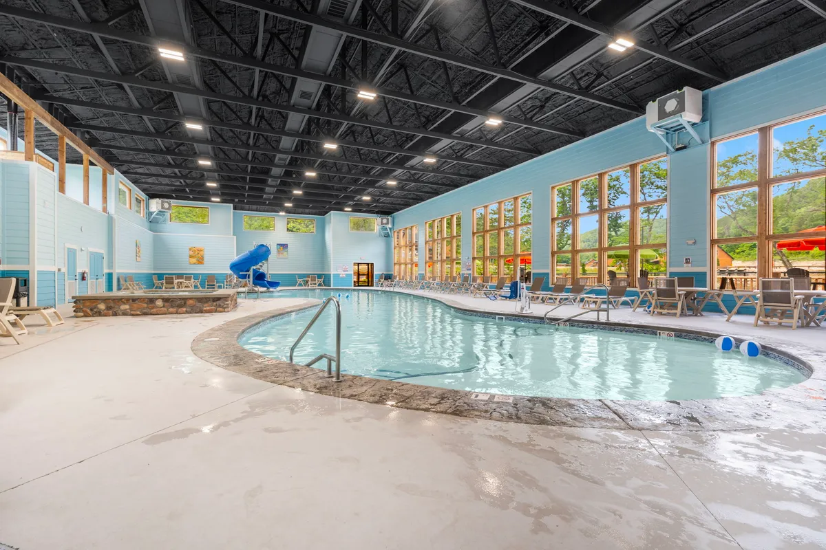A spacious, light-filled indoor pool area featuring a waterslide, expansive windows, and multiple lounge chairs. The pool is inviting, with vibrant blue water and plenty of space for families to relax or play. Natural light pours in from the windows, creating an energizing and cheerful atmosphere.