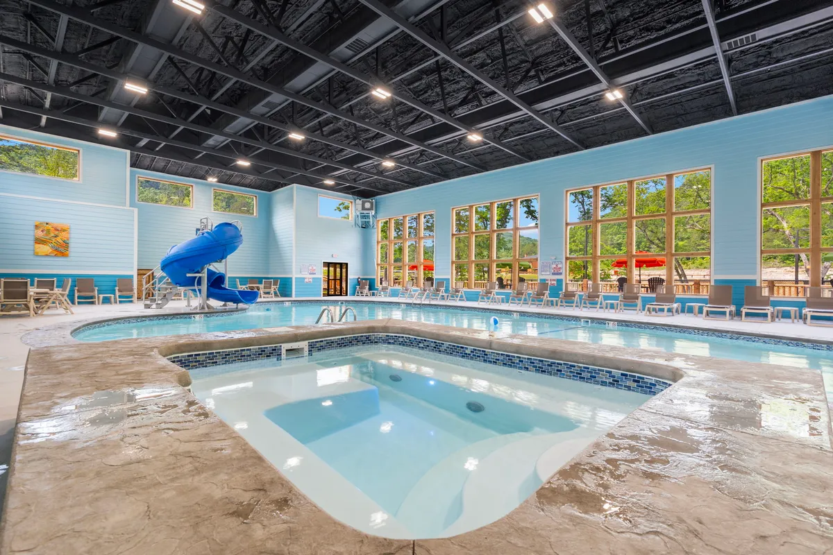 An inviting indoor swimming pool area, complete with a water slide, spa, and ample lounge seating. Large windows let in natural light, and families or groups are seen enjoying the water together, creating a lively and playful atmosphere.