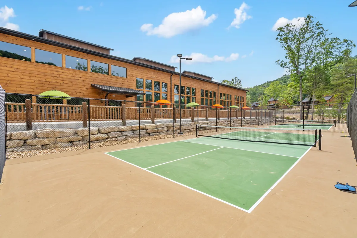 Well-maintained tennis courts next to a wooden building, shaded by green umbrellas. The courts are set in a natural outdoor space, ideal for friendly matches, group activities, or simply enjoying the fresh air.