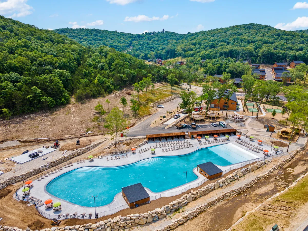 An inviting outdoor pool area surrounded by lush green hills, a wooden deck, and colorful umbrellas. Lounge chairs are arranged for sunbathing or relaxing, and the setting conveys a joyful and social vibe ideal for warm weather activities.