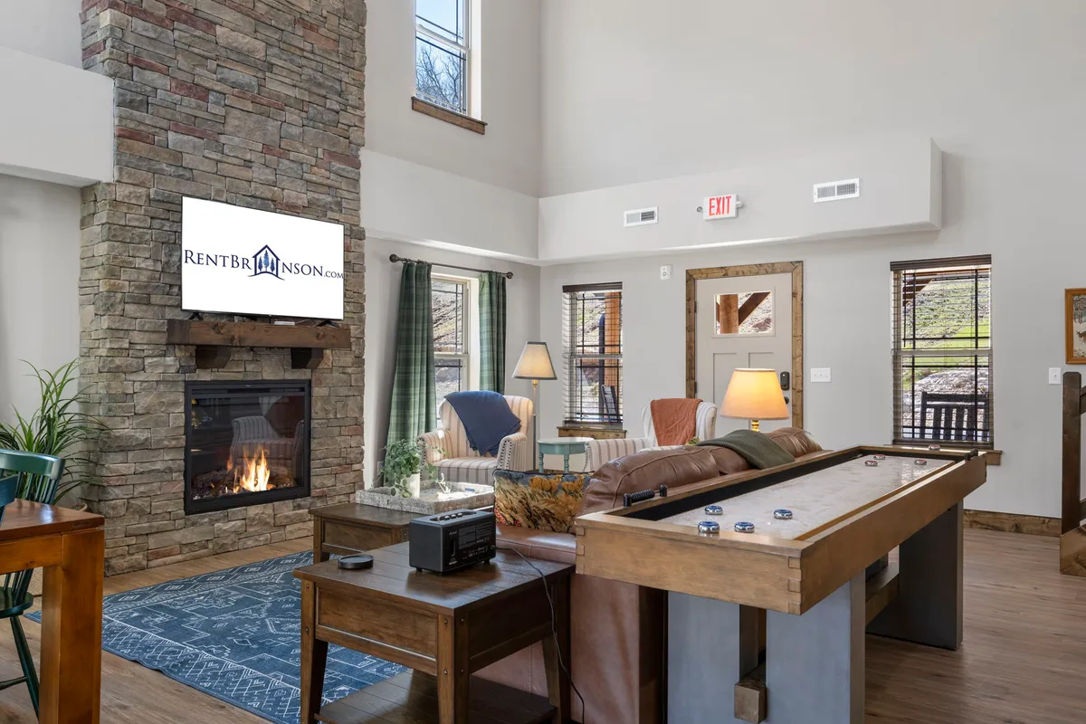 The image showcases a warm, inviting living area featuring a crackling fireplace, a plush sofa, a sleek flat-screen TV, and a pool table—all framed by large windows that offer serene woodland views. The scene is set for relaxation, family fun, and cozy gatherings, with natural light pouring in to highlight the open-concept design.